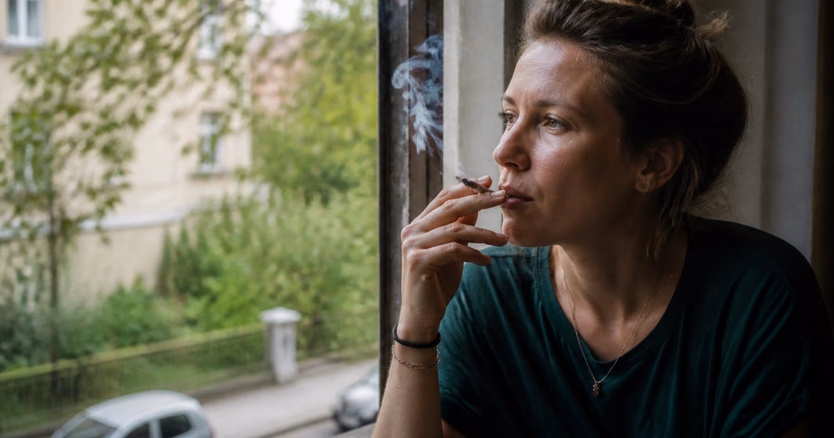 Nő jointot szív az ablaknál, miközben egy fákkal szegélyezett utcára néz