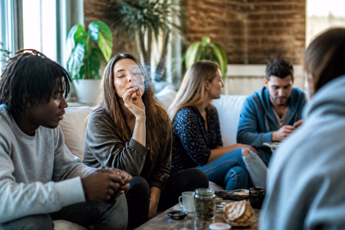 Fiatal felnőttek egy kanapén, egy nő füves cigarettát szív, az asztalon kannabiszos befőttesüveg